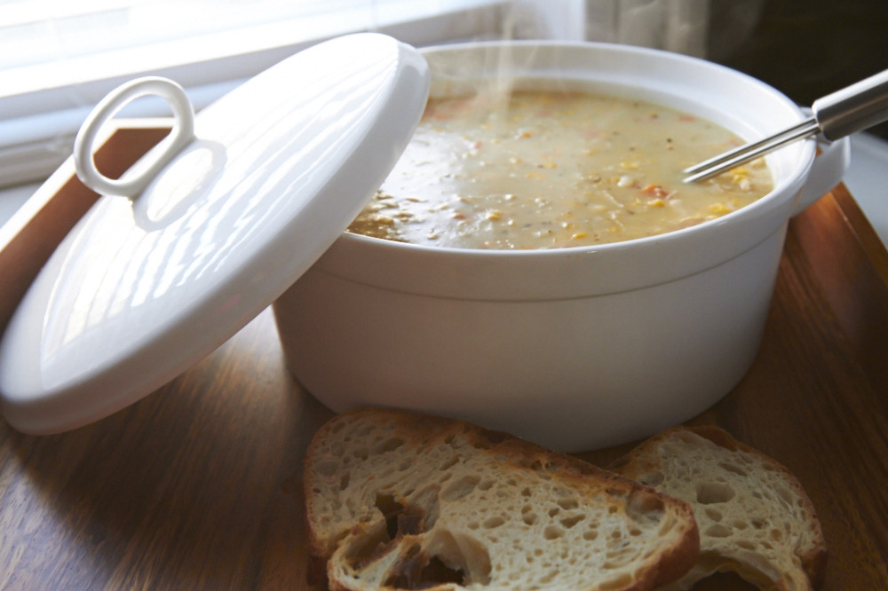 A steaming pot of chicken pot pie soup sits on a wooden tray next to two slices of crusty bread. 