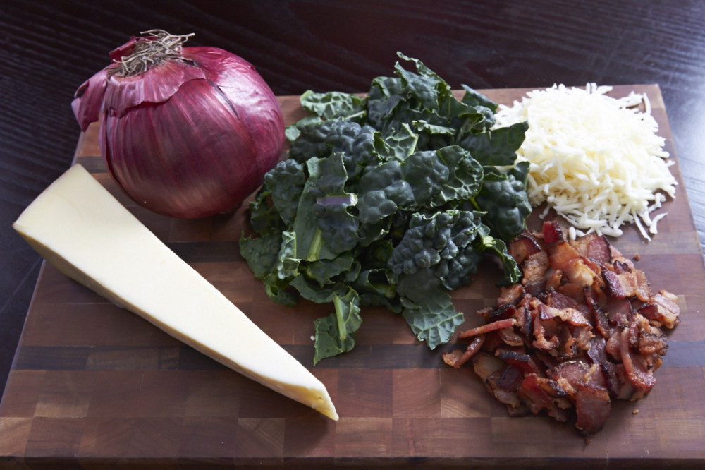 A wooden cutting board with an onion, a block of cheese, fresh kale, cooked bacon and shredded cheese.