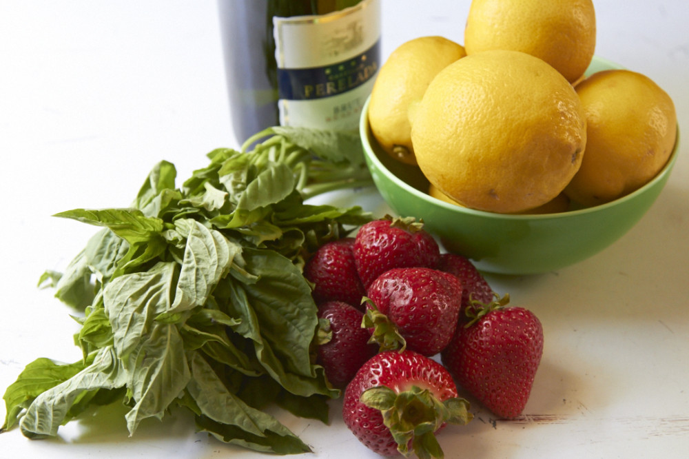 A bowl of lemons, some fresh strawberries, a bunch of fresh basil and a bottle of Prosecco on a white surface.