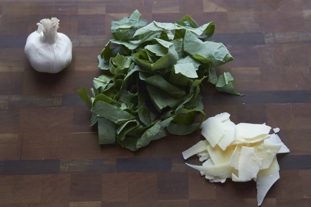 Fresh kale, a head of garlic, and shredded parmesan cheese on a wooden cutting board.