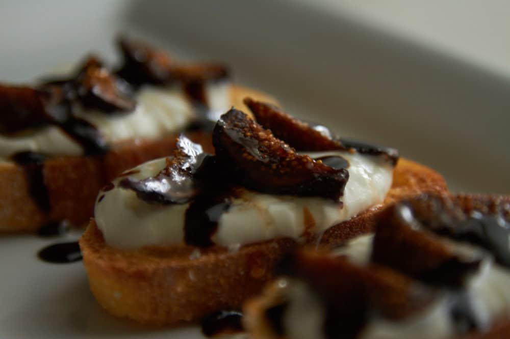 Crostini with whipped goat cheese, figs and balsamic glaze on a white plate. 