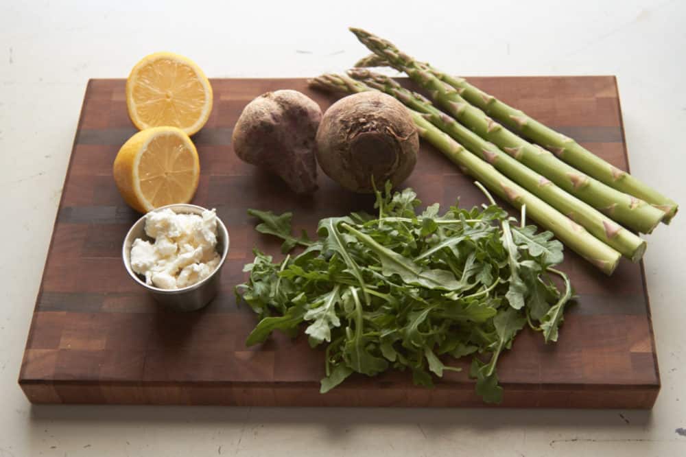 Lemon halves, beets, asparagus, arugula and goat cheese on a wooden cutting board.
