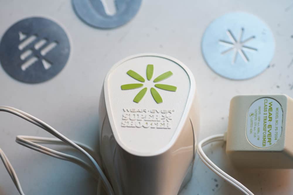 An electric cookie press with wording that says Wear Ever Super Shooter, surrounded by small metal discs for making cookies of various shapes. 