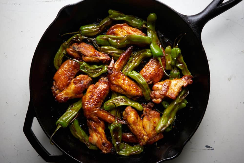 Honey-Harissa chicken wings with shishito peppers in a cast iron skillet displayed on a white surface. 