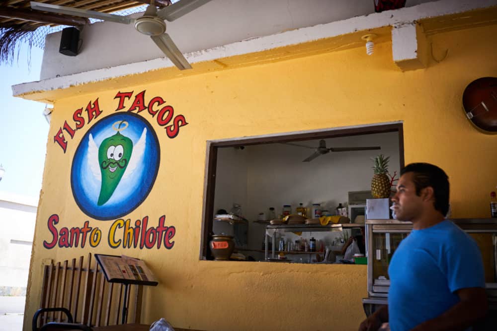 Fish Tacos Santo Chilote restaurant in Todos Santos Mexico. A yellow wall with a green chile logo to the left of a food pickup window. A waiter in a blue shirt stands to the right of the window. 
