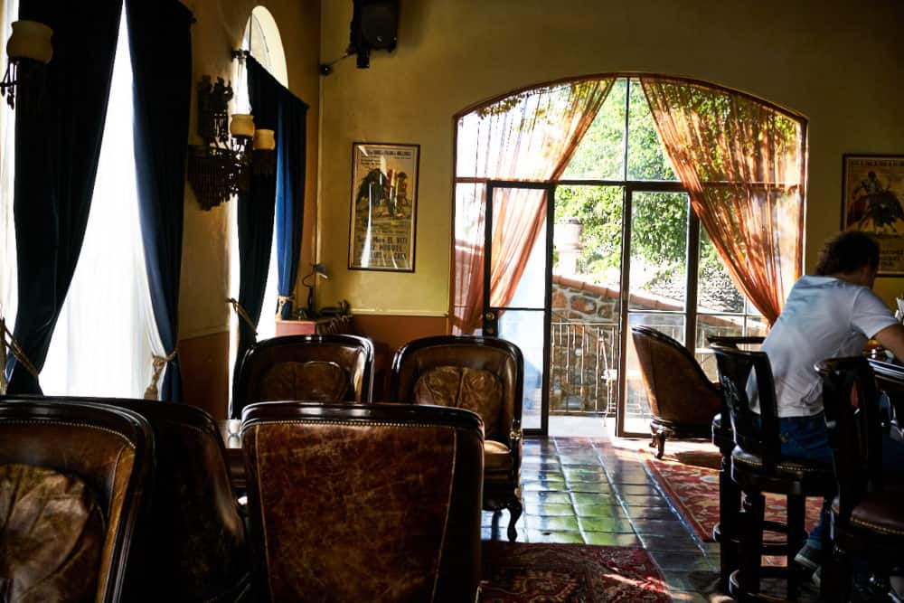 The bar at the Todos Santos Inn. A high-ceilinged room with yellow walls, stone tile floors, big leather arm chairs and large arched windows with blue curtains. the far wall has large glass doors that open onto a courtyard. 
