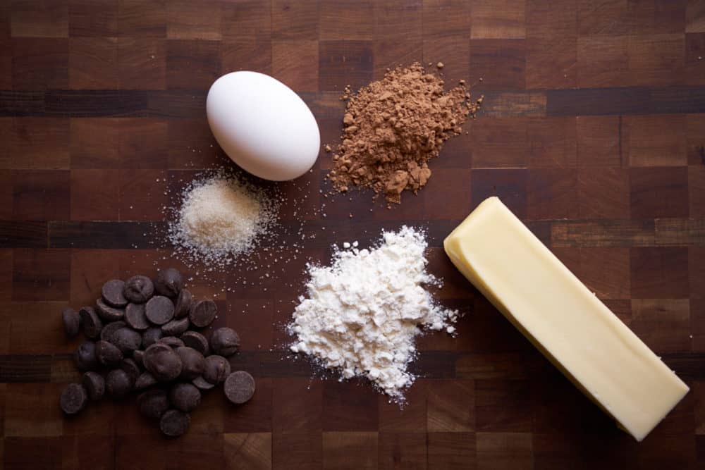 Chocolate chips, flour, cocoa powder, sugar, an egg, and a stick of butter displayed on a wooden cutting board.