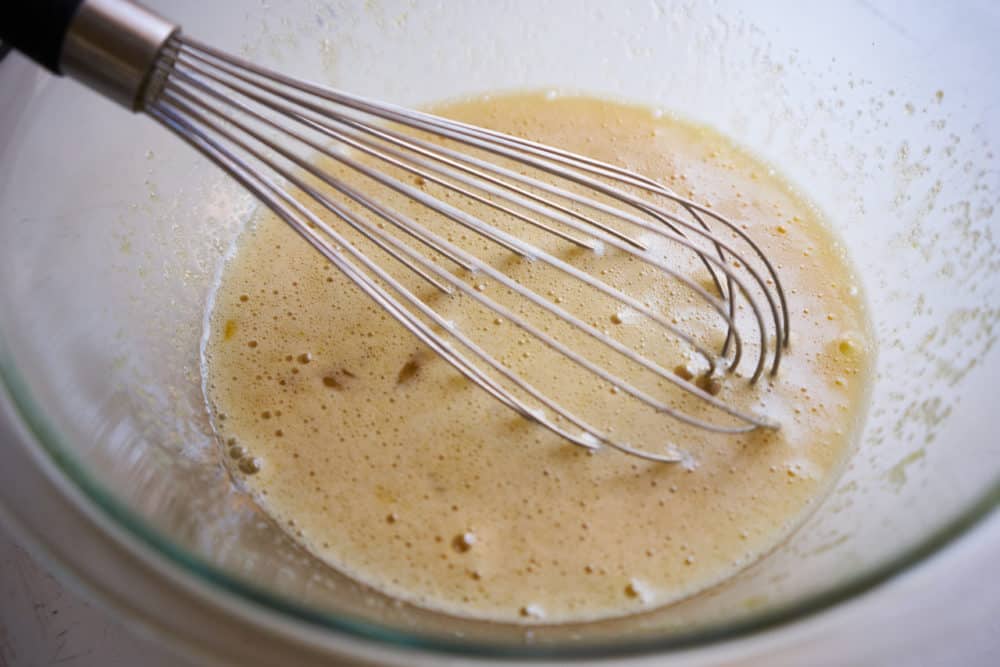 A whisk in a glass bowl containing eggs, sugar and vanilla that have been whisked together into a frothy mixture.