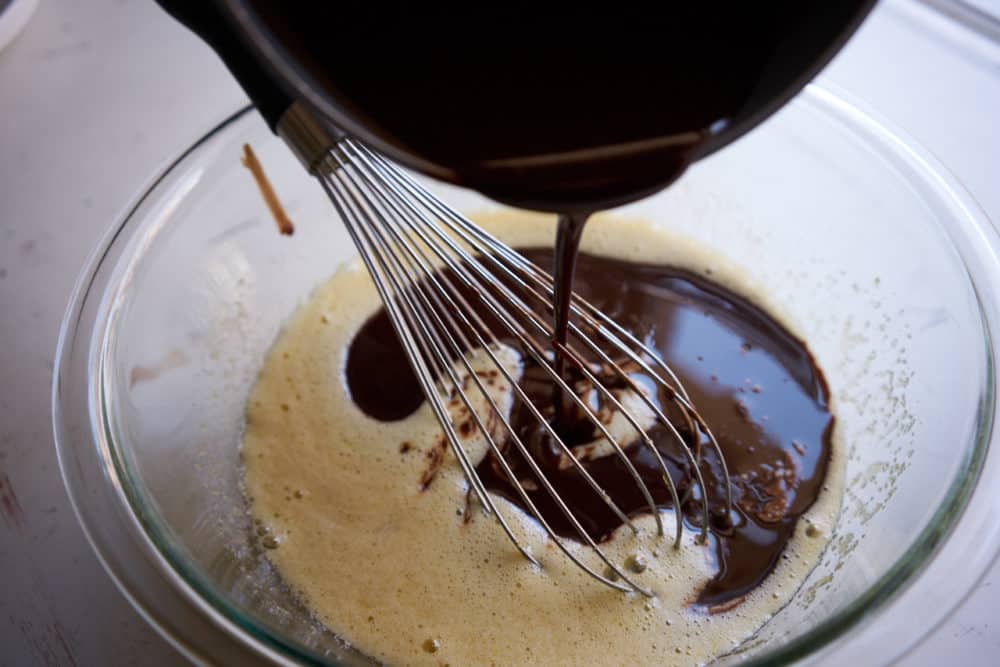 Chocolate is poured from a small sauce pan into a glass bowl with a whisk in it containing an egg and sugar mixture.
