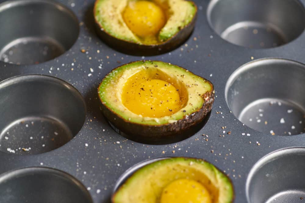Raw eggs inside avocado halves sitting inside the wells of a muffin pan.