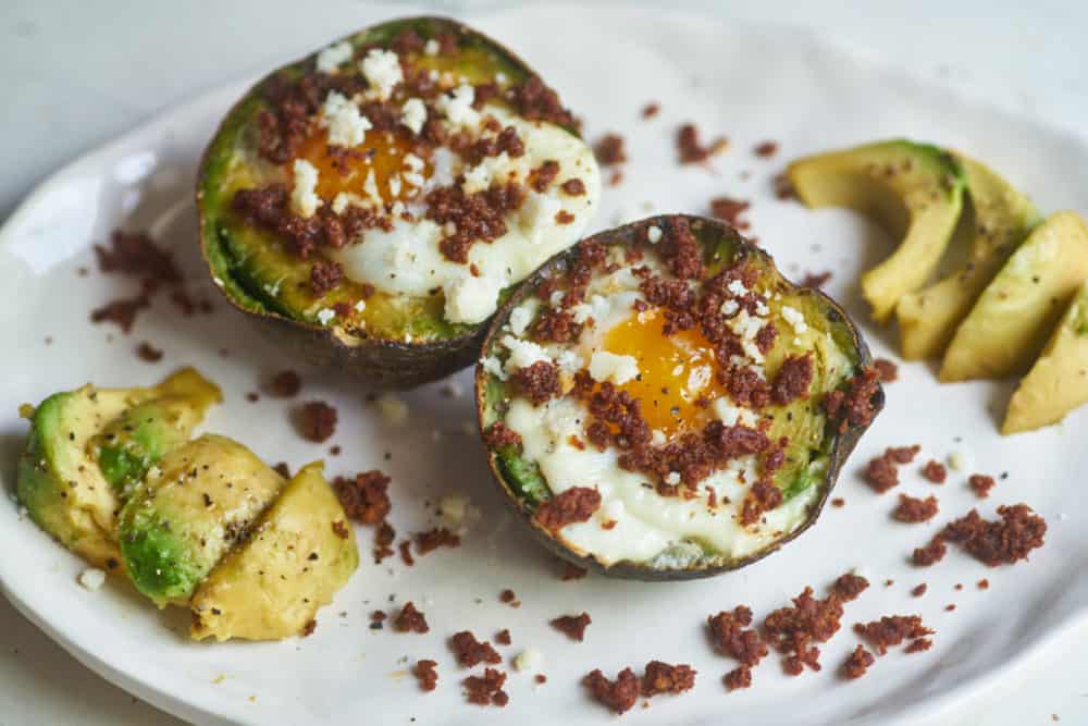 Two eggs baked in an avocado half topped with chorizo and queso fresco displayed on a white plate, surrounded by bits of chorizo and avocado slices.