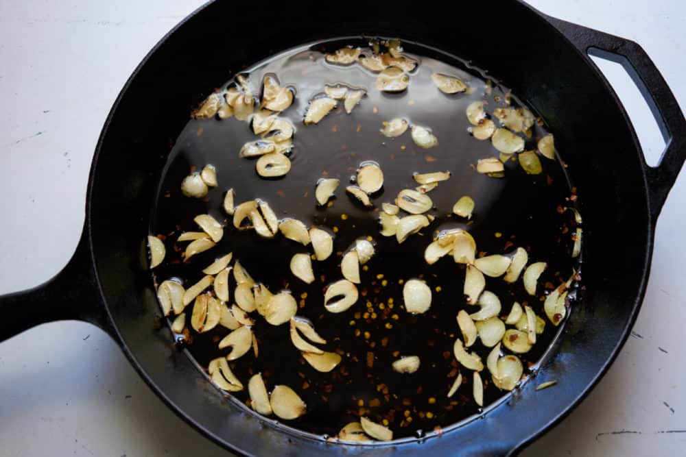 A cast iron pan is filled with sliced garlic and crushed red pepper that have been simmered in olive oil.