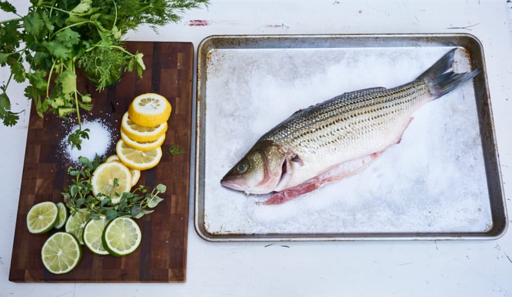 Whole fish on a bed of salt on a sheet pan next to a cutting board with fresh herbs and sliced lemons and limes.