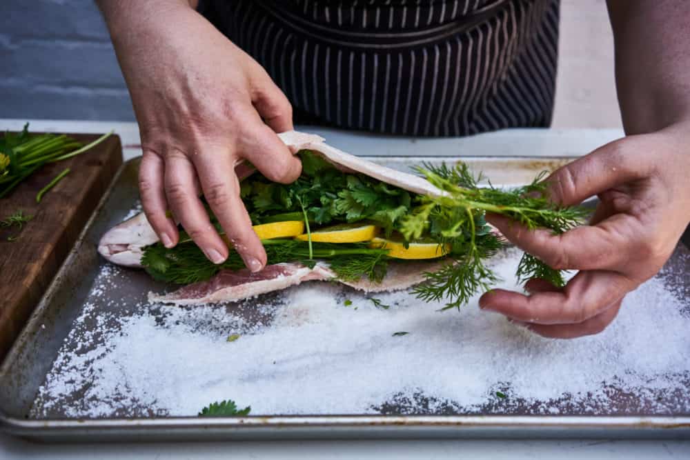 A woman stuffs a whole fish with lemon, lime and fresh herbs. The fish is laying on a bed of salt on a sheet pan.