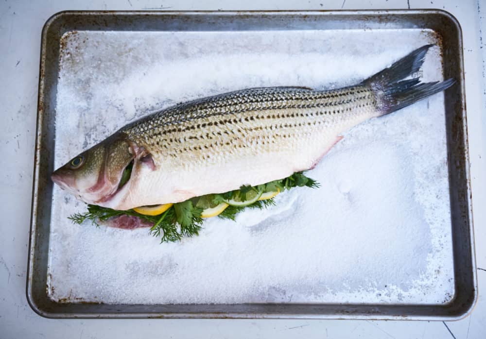 A whole striped bass stuffed with herbs and citrus fruit sits on a bed of salt on a silver sheet pan. 
