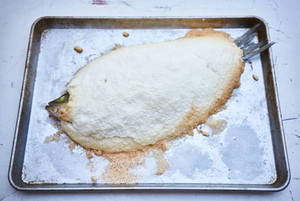  A whole fish that has been baked in a salt crust is shown on a silver sheet pan. The edges of the salt are browned. 