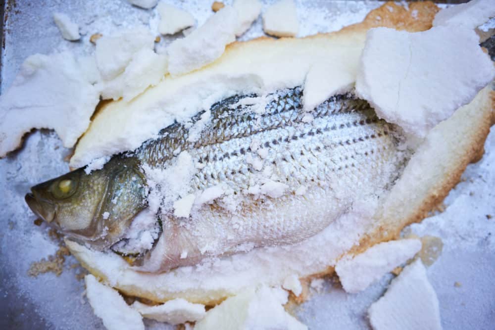 Whole fish baked in salt on a silver tray surrounded by chunks of salt crust.