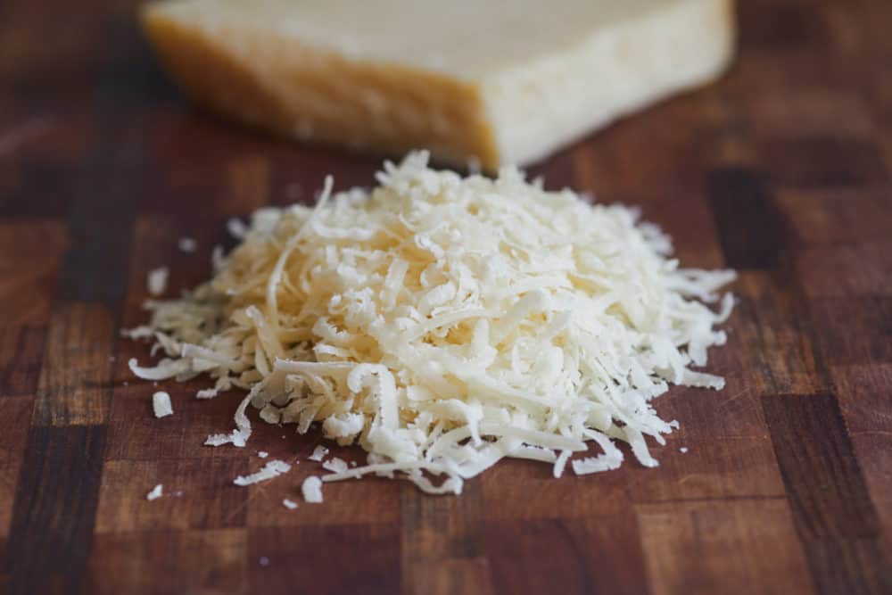 Shredded parmigialno reggiano sits in the foreground on a wooden cutting board, with a block of the cheese in the background.