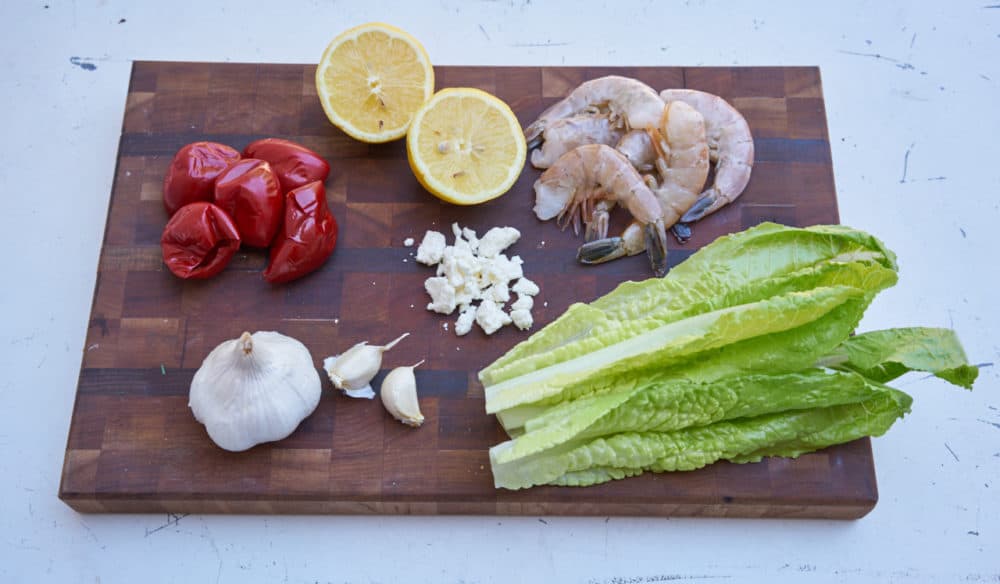 Romaine lettuce leaves, feta cheese, garlic, cherry peppers, lemon and raw shrimp displayed on a wooden cutting board. 