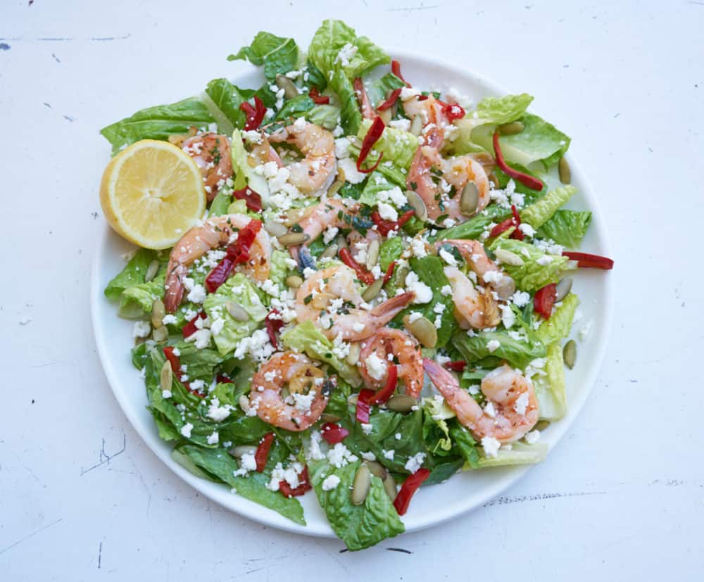 Large round plate of salad with shrimp, romaine lettuce, cherry peppers, pepitas and feta cheese, garnished with a half a lemon.