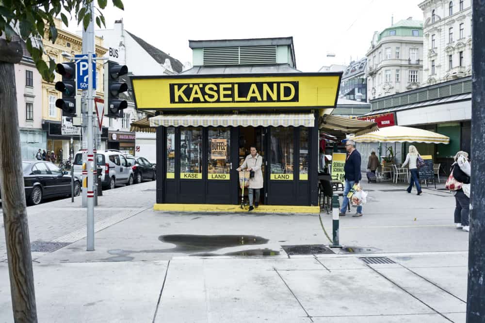 Käseland shop at Rochusmarkt Vienna. A yellow and black building, a woman is exiting the front door with a scooter.