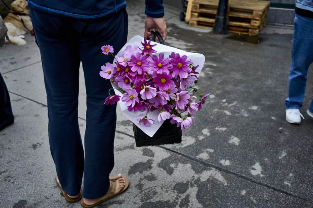 A man wearing blue pants and tan flip flops shown from the waist down holds a basket of purple flowers at Rochusmarkt in Vienna.