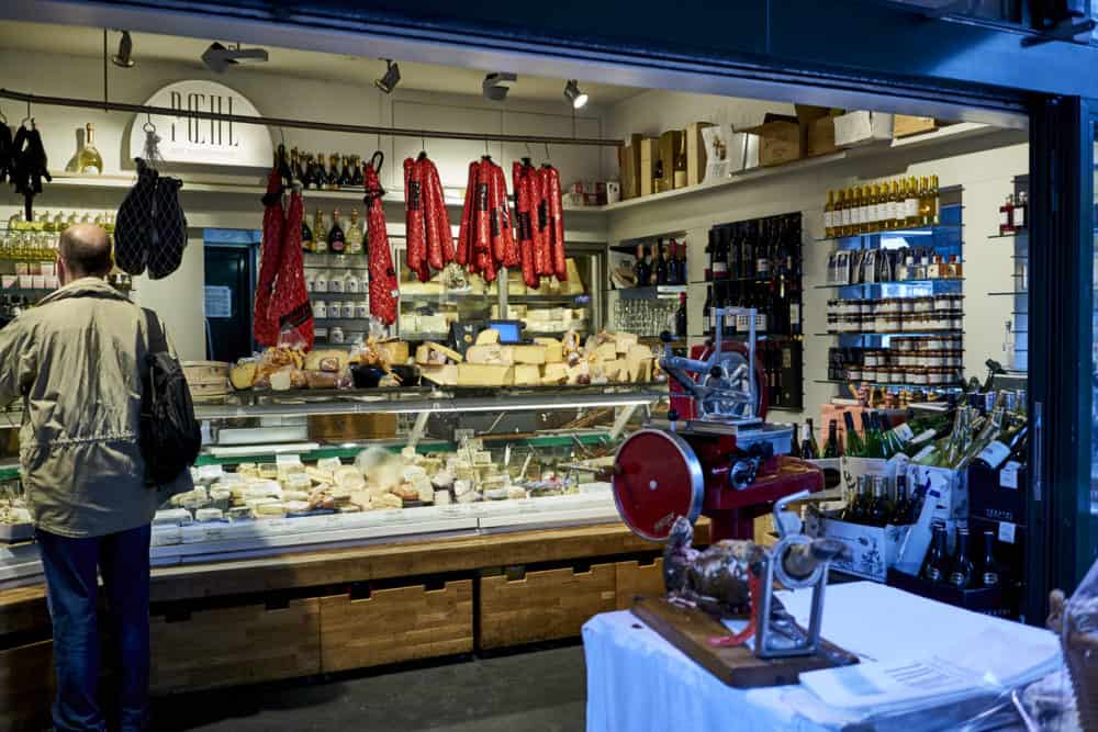 A cheese and charcuterie stand at Naschmarkt in Vienna. A deli case holds a variety of cheeses and cured salamis hang from the ceiling. A meat grinder is in the foreground.