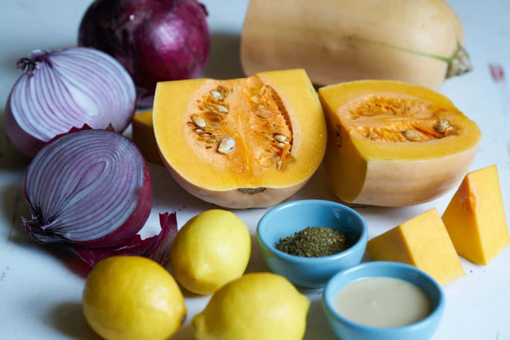 A butternut squash cut in half is surrounded by a halved red onion, whole lemons, and small blue bowls filled with za'atar and tahini.