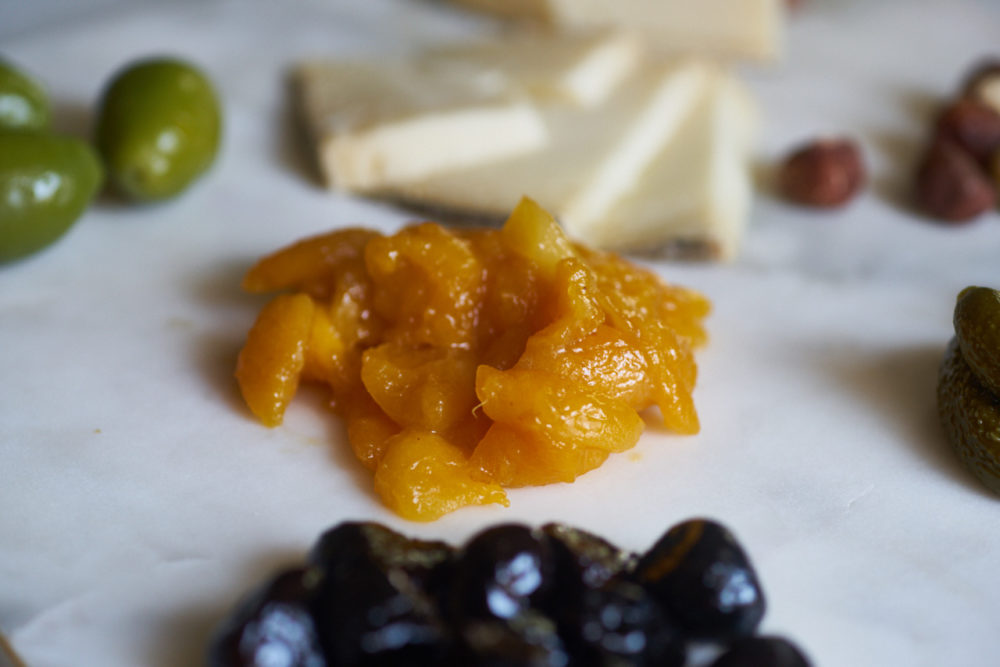A variety of ingredients for a cheese plate including mostarda, olives, pickes, hazelnuts and cheese, on a marble surface.