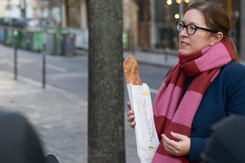 A tour guide from Paris by Mouth stands on the sidewalk in the Marais neighborhood in Paris. She holds a baguette, she is wearing a blue coat and a red and pink striped scarf. 