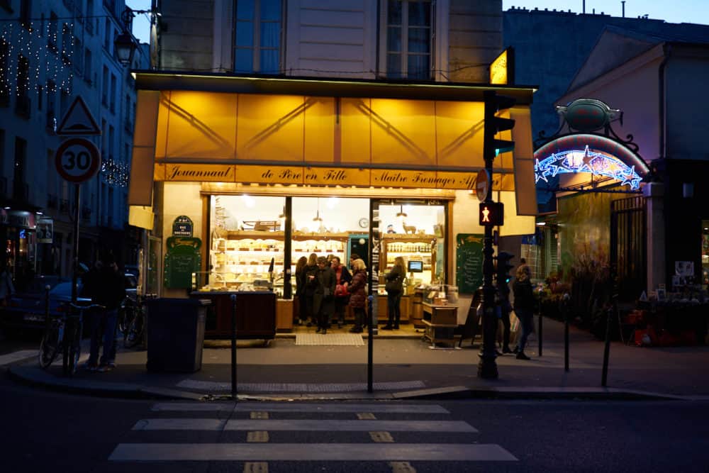 The exterior of Fromagerie Jouannault, a cheese shop in the Marais neighborhood of Paris. The awnings are yellow, a cross walk is in the foreground. 
