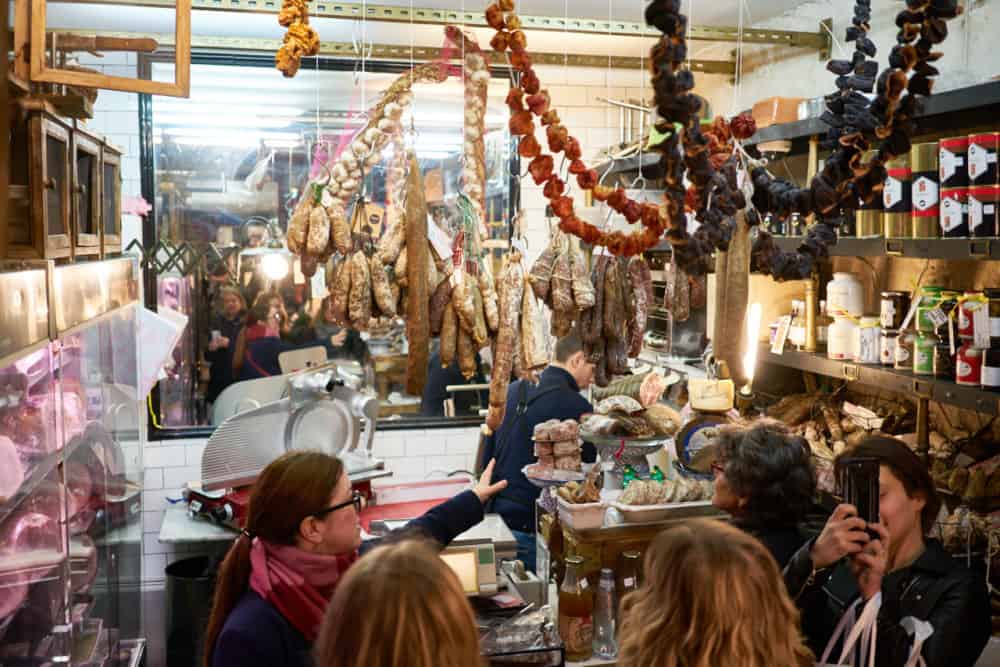 The interior of Caractère de Cochon, a charcuterie in the Marais neighborhood in Paris. Sausages and dried peppers are hanging from the ceiling. A tour guide and tour group is in front of the counter as a man slices charcuterie behind it. 