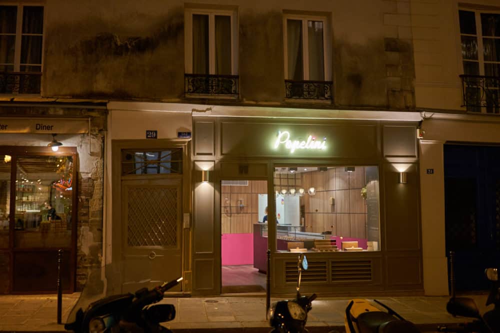 The exterior of Popelini in the Marais neighborhood of Paris. The shop sells small cream puffs in a variety of flavors. A white neon sign that says Popelini is lit over the entrance. 