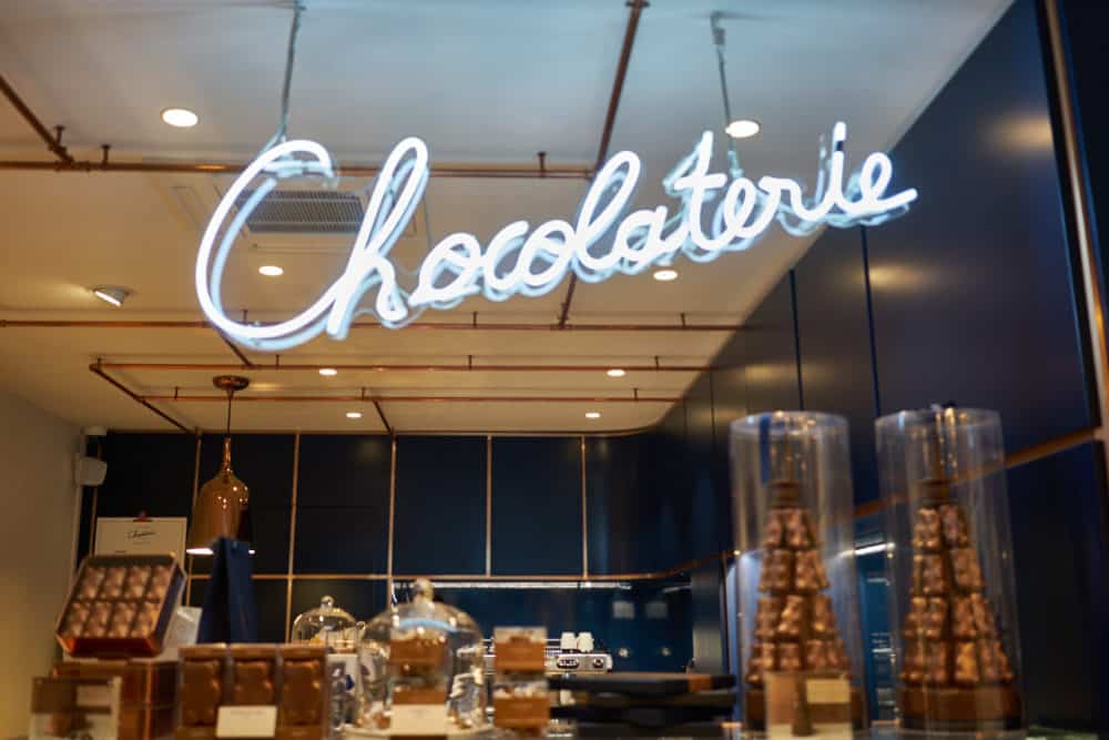 Inside La Chocolaterie Cyril Lignac in Paris. A white neon sign that says "Chocolaterie" hangs over a counter with pastries and chocolate. The interior walls are dark blue. 