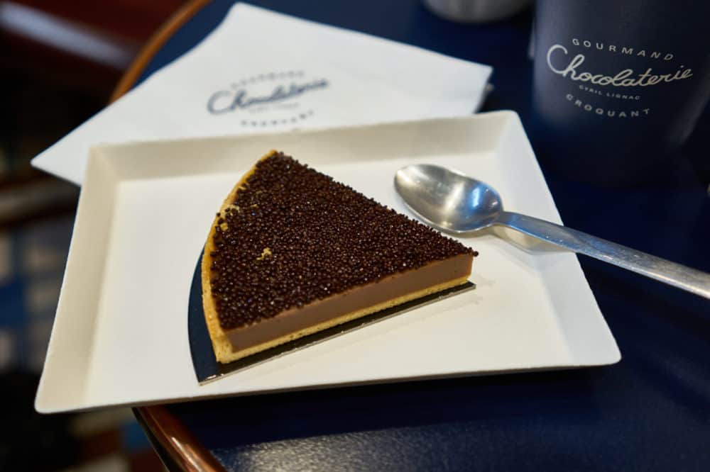 A chocolate hazelnut tart on a white plate sits on a blue table at chocolaterie Cyril Lignac in Paris. A spoon is resting on a plate and a napkin with the chocolaterie's logo is above the plate. 