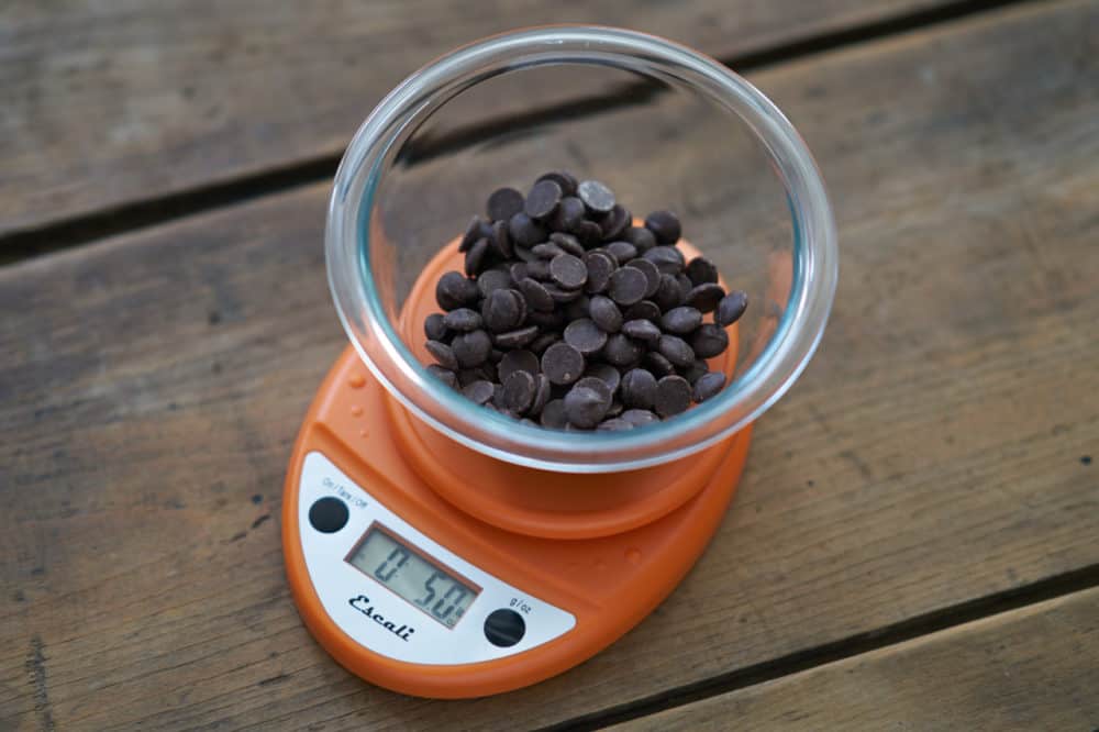 An orange kitchen scale weighs a glass bowl filled with dark chocolate chips.