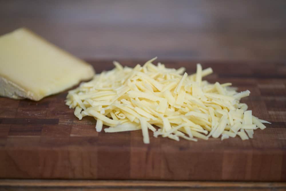 A block of comté cheese and some shredded comté cheese on a wooden cutting board sitting on a wooden table.