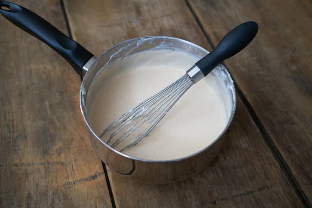 A saucepan filled with bechamel sauce that has paprika added to it sits on a wooden surface. A whisk is in the pot leaning to the right. 