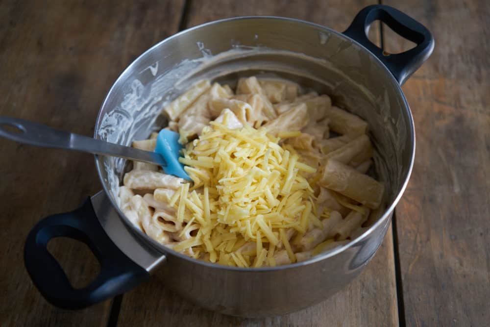 A deep pan filled with cooked pasta coated with bechamel sauce, grated comté cheese has been added but not yet mixed in. A spatula sits in the pan, leaning to the left. 