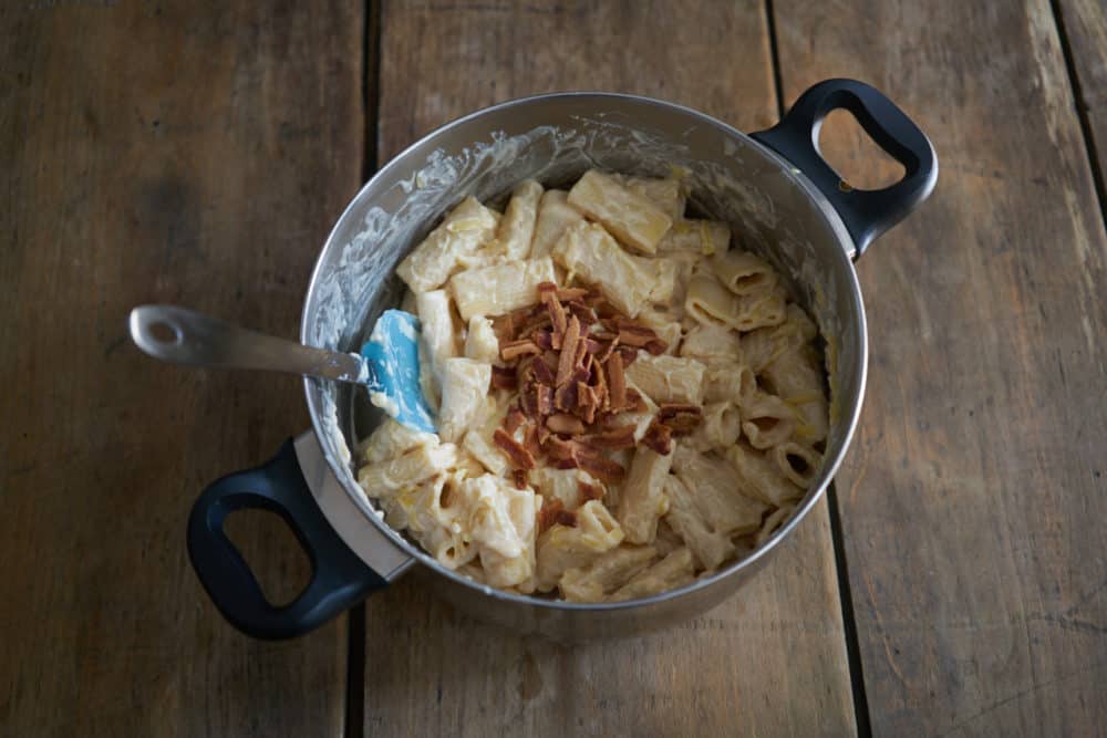 A deep pan filled with cooked pasta with a cheesy cream sauce. Crumbled bacon is in the center of the pan. A spatula sits in the pan, leaning to the left. 