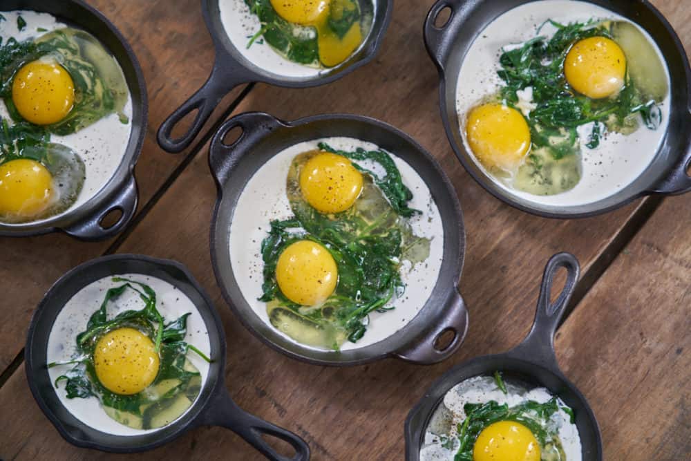 Several small cast-iron skillets and serving dishes containing a small amount of cooked spinach, raw eggs, and some heavy cream are displayed on a wooden surface. 