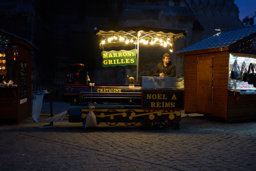 A roasted chestnut cart at the Christmas market at Reims cathedral in France. The cart is decorated like a steam train engine in green and yellow. Signs say Marrons grilles, Noël á Reims.