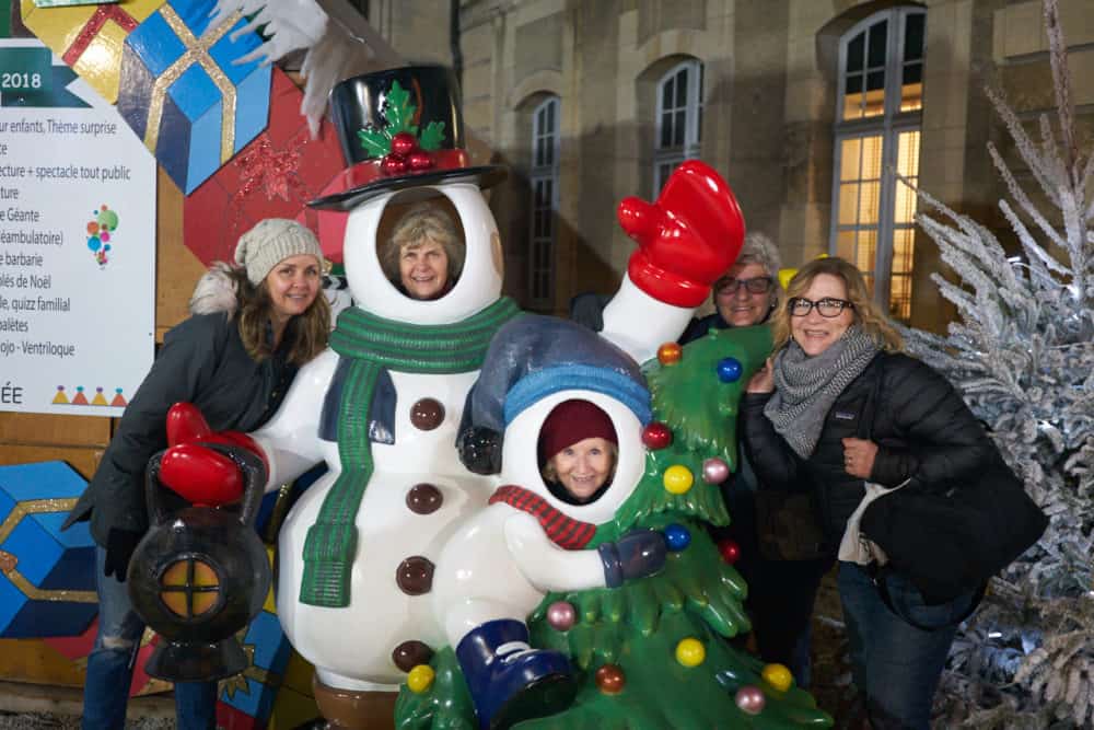 Five women are posing with a Christmas snowman statue at the Christmas market in Reims, France. Two of the women have their faces where the snowman's faces should be. They are all laughing and smiling.