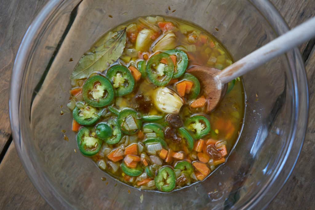 A vinegar sauce containing cooked carrots, onions and whole garlic cloves, with golden raisins and sliced fresh jalapeños in a glass bowl with a wooden spoon.