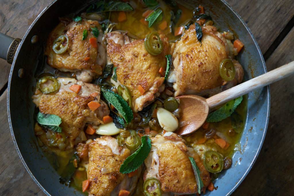 A skillet with chicken escabeche sits on a wooden surface. A wooden spoon is in the pan on the right side. Garlic, golden raisins and jalapeños can be seen in the escabeche sauce, and the dish is garnished with fresh mint.