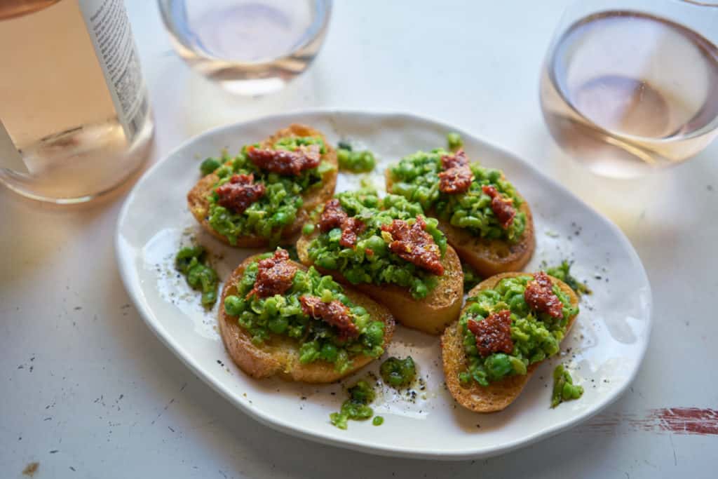 A plate of crostini with peas, mint and nduja along side a wine bottle and two glasses filled with rosé wine.