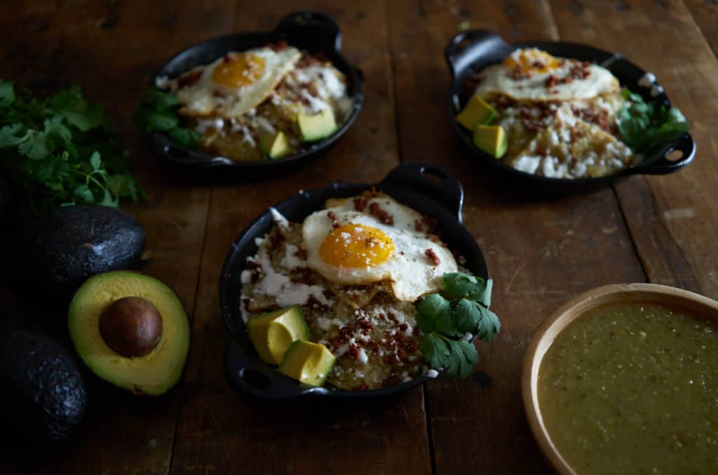 Three small cast iron dishes of chilaquiles verdes topped with a fried egg sit on a wooden table.