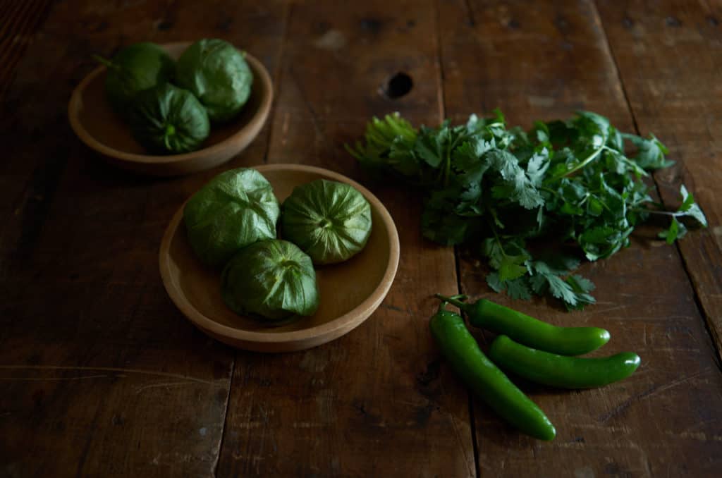 Tomatillos in wooden bowls, serrano chiles, and cilantro on a wooden table. 