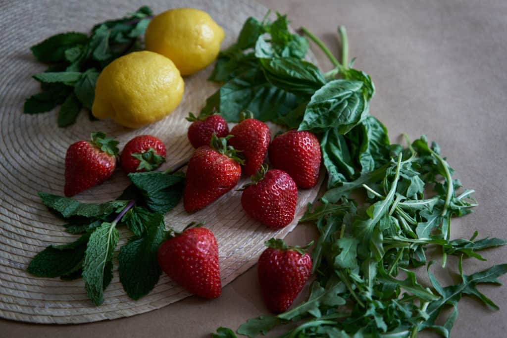 Strawberries, lemons, mint, basil, and arugula on a woven placemat.