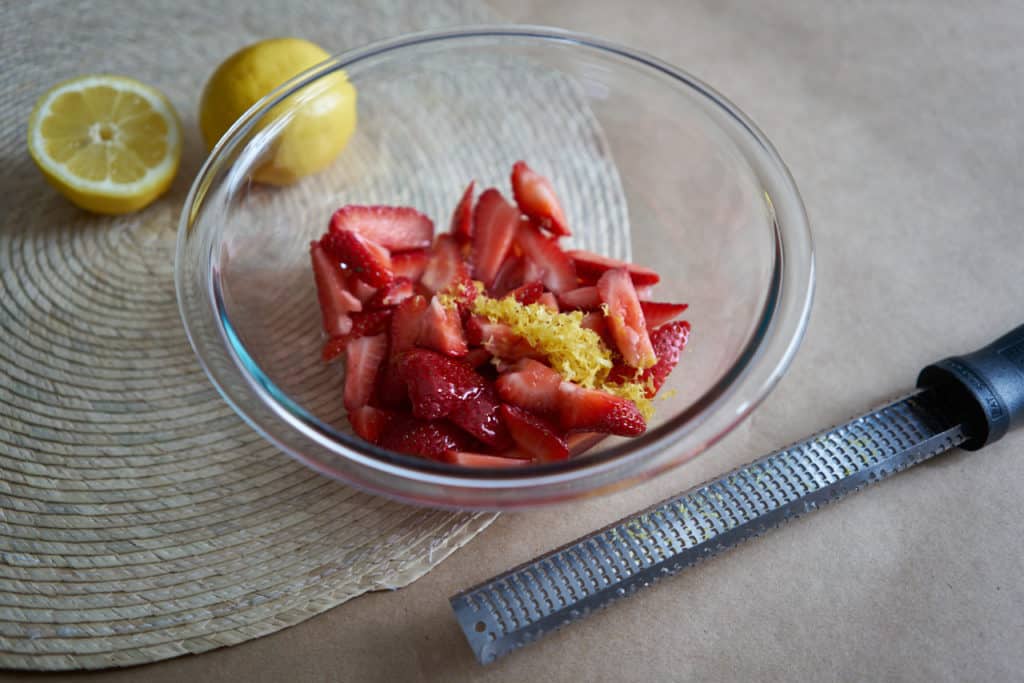 A glass bowl filled with sliced strawberries and lemon zest sits on a woven placemat. A zester is in the foreground and lemons are in the background.